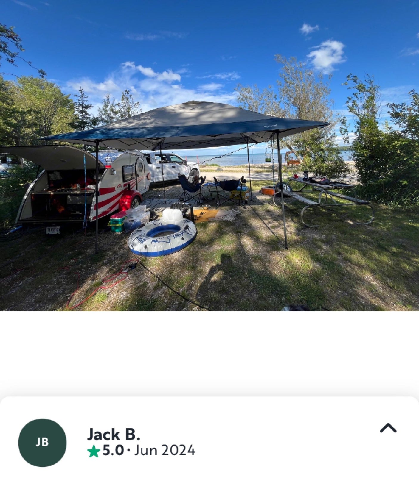 Jack B.'s 5-star review: An RV rental teardrop trailer campsite setup by the water with a large canopy, truck, and gear.