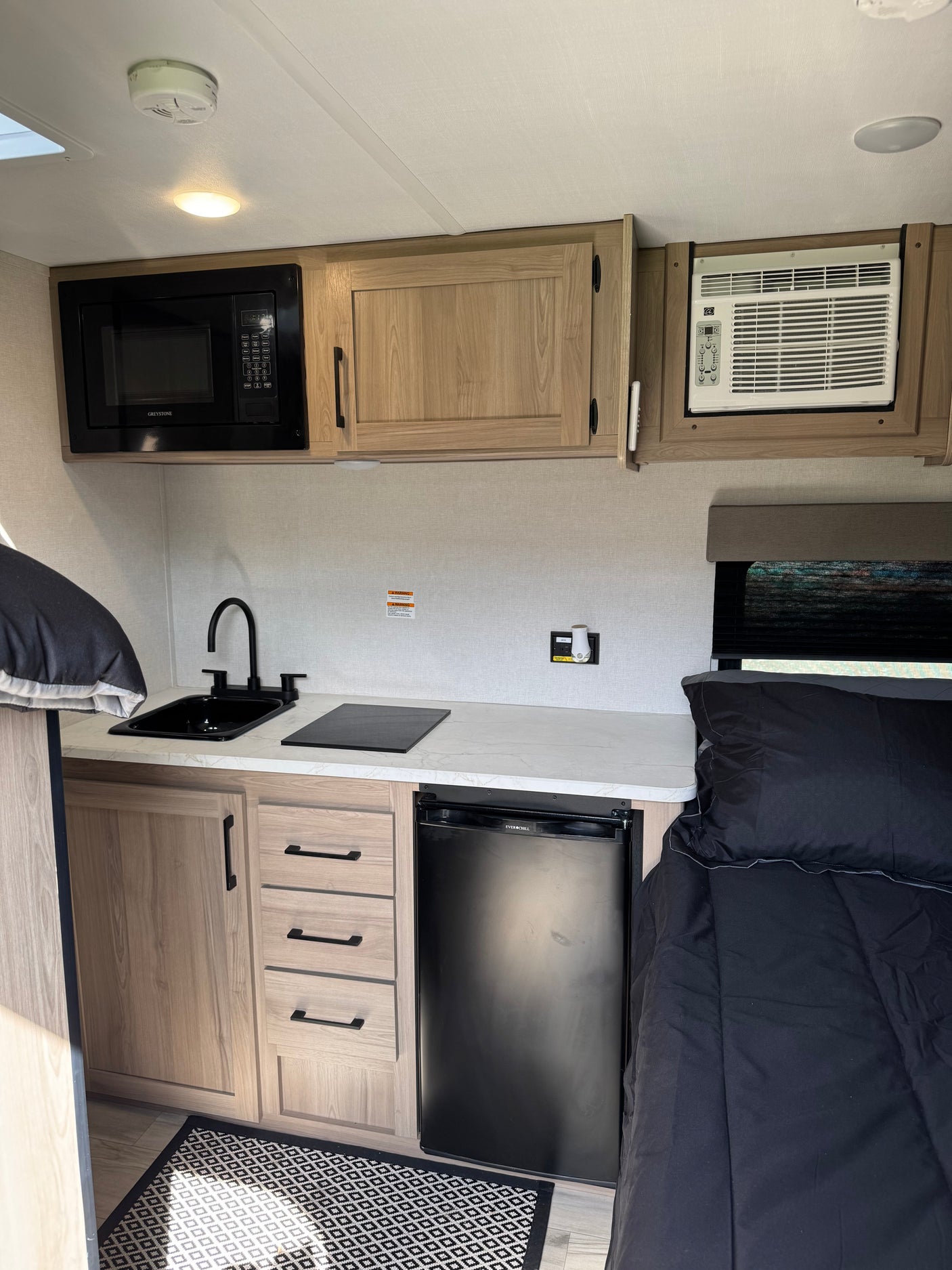 Inside a modern RV rental: compact kitchen with microwave, sink, fridge, and cozy bed area.