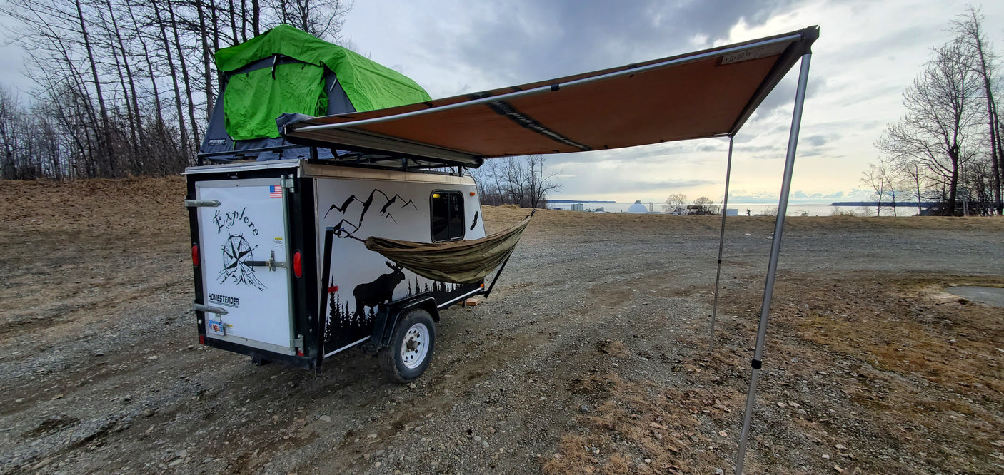 Overland trailer with rooftop tent, hammock, and awning, set up for camping near a body of water—ideal for an RV rental adventure.
