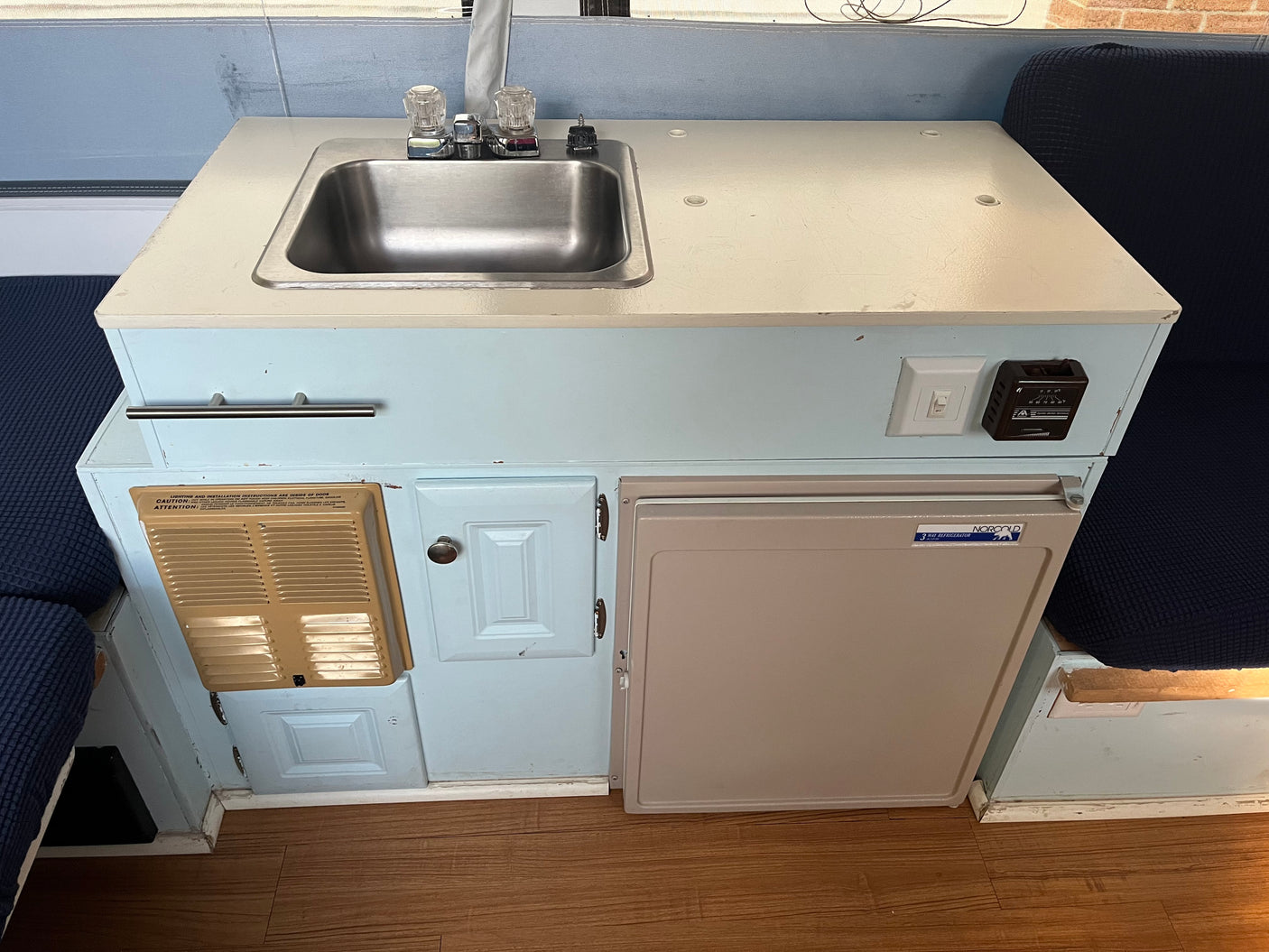 Compact RV rental kitchenette featuring a stainless steel sink, heater vent, storage cabinet, and Norcold refrigerator.