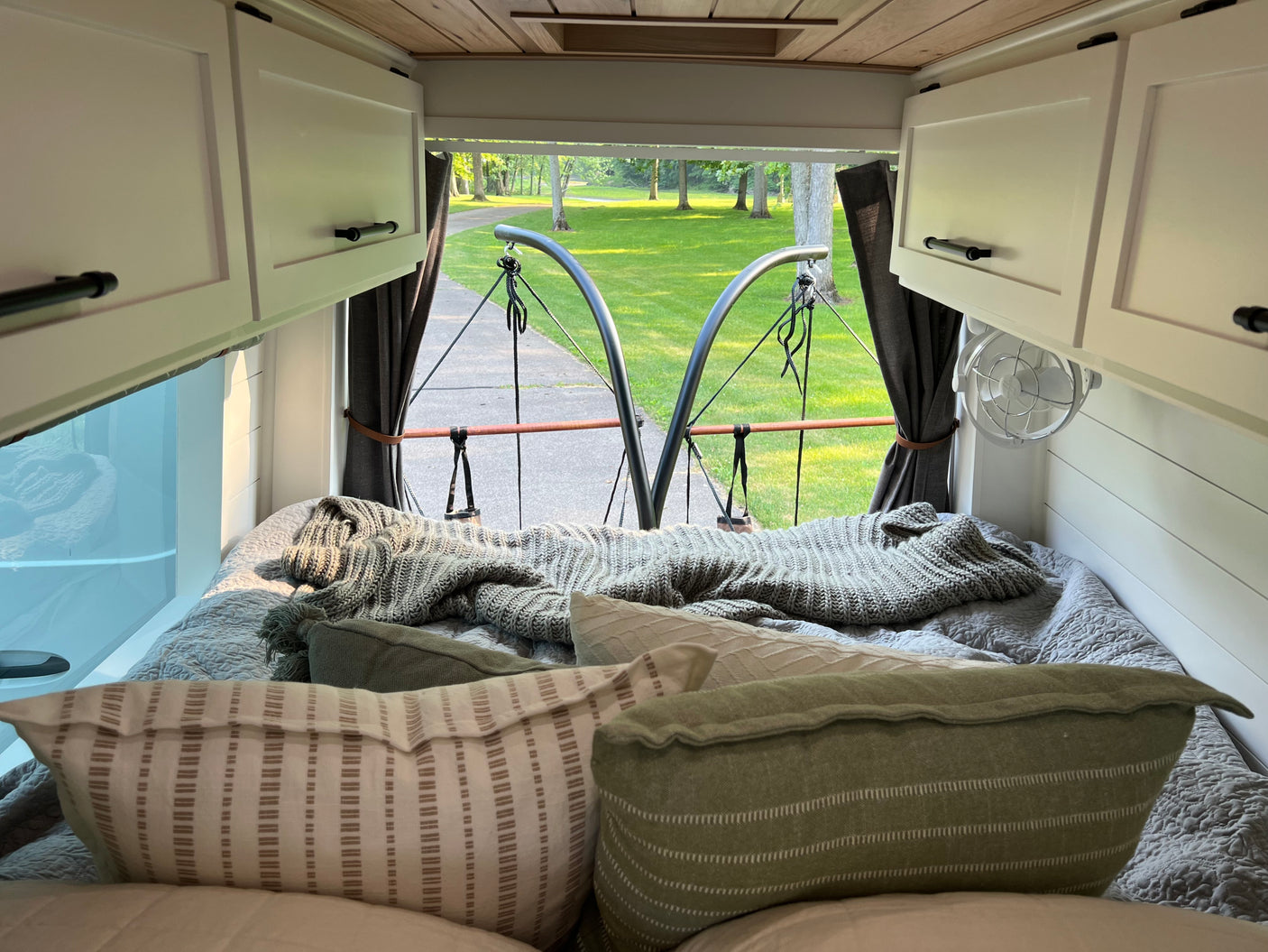 Relax in a cozy RV rental: This image shows a comfortable bed inside an RV, looking out the back to a park-like view with trees and a paved path.