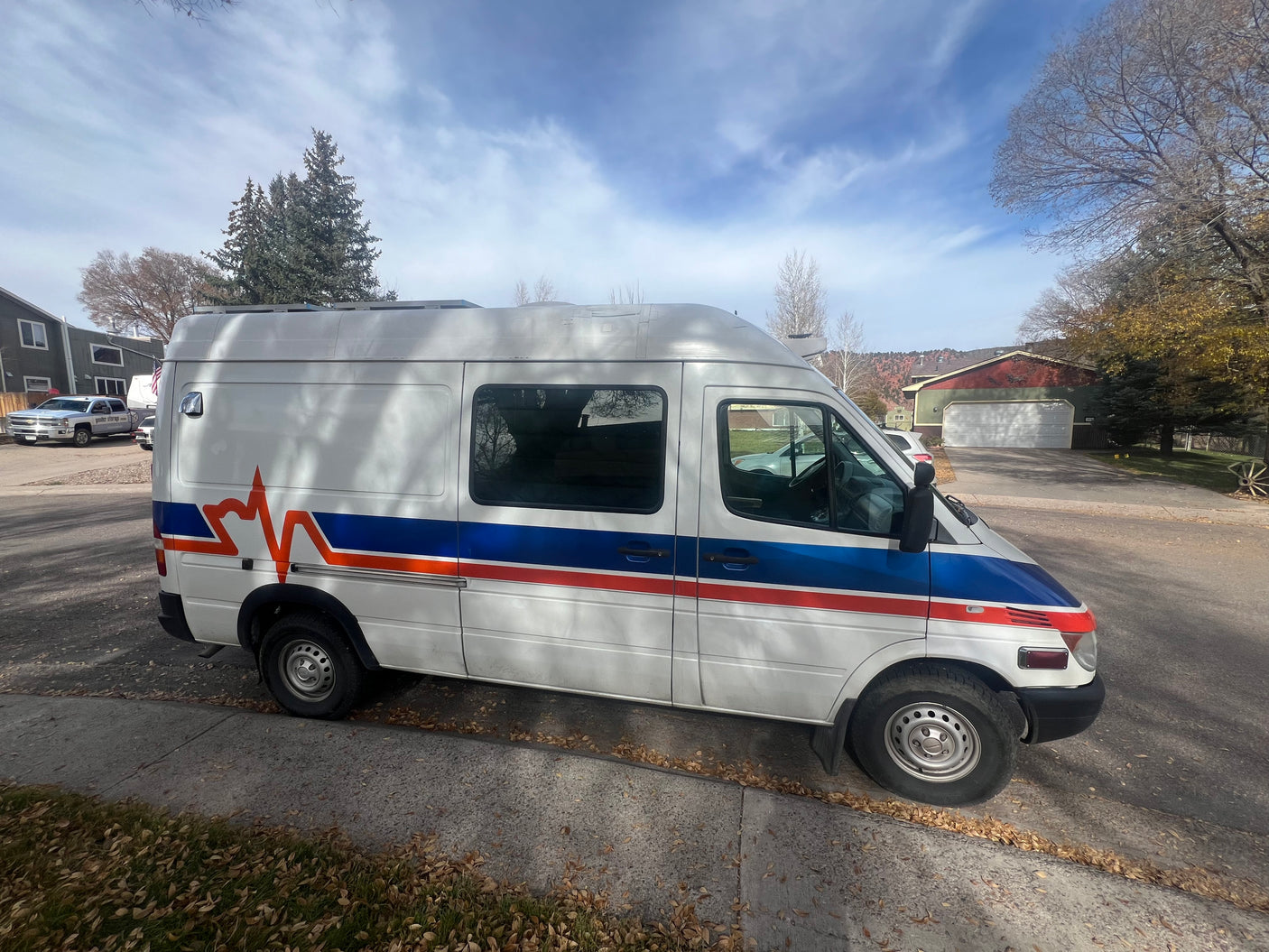 White Sprinter-style van with blue/red ECG stripe, possibly a custom rv rental, parked on a fall street.