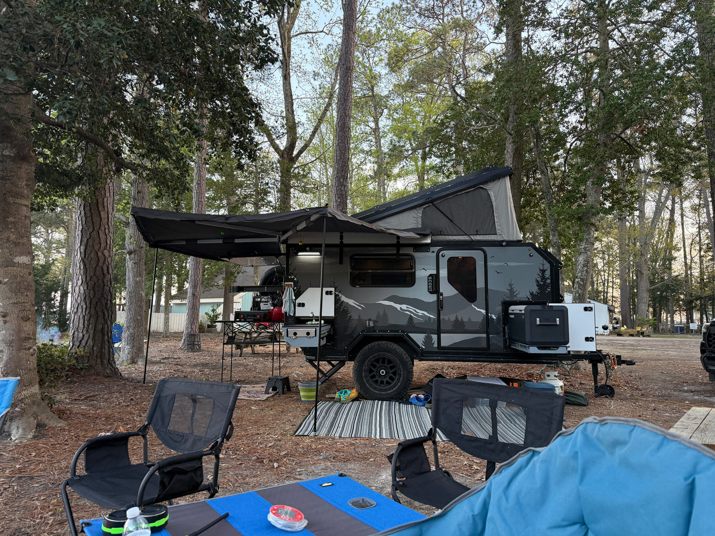 Overland rv rental camper with outdoor kitchen, pop-top tent, and awning setup at a wooded campsite with chairs.