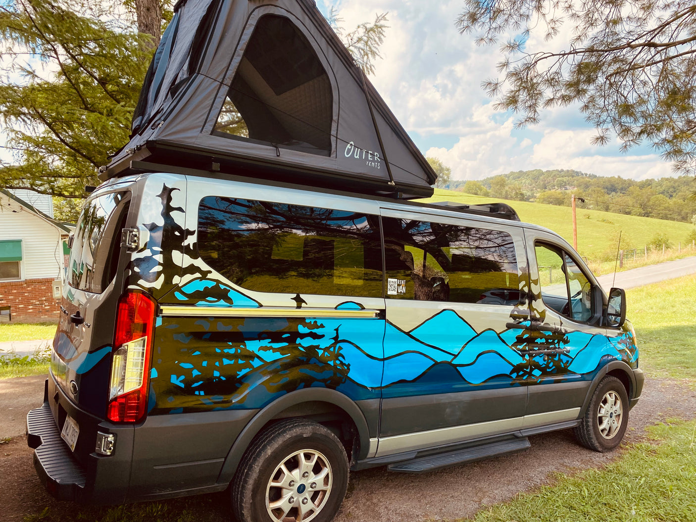 A custom camper van with a mountain/forest wrap & open rooftop tent, perfect for an RV rental in scenic outdoors.