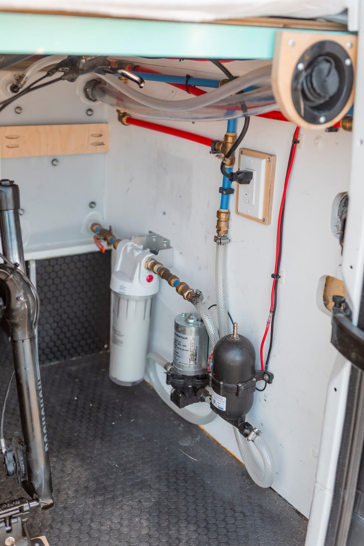 View of an RV rental's internal water system: pump, filter, and PEX plumbing. A bike fork is visible on the floor.
