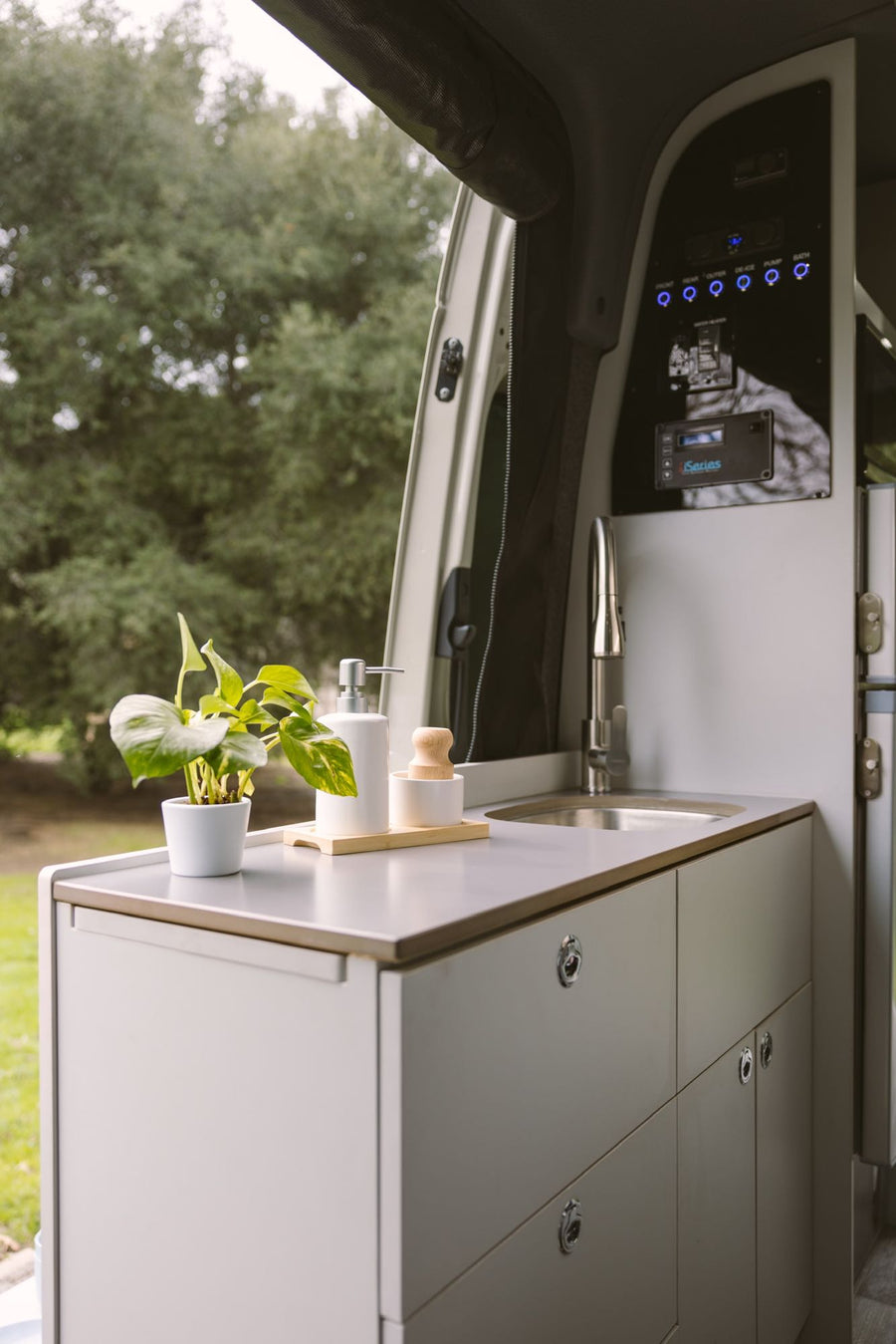 An RV rental's modern interior kitchen with a sink, faucet, potted plant, and control panel, offering a view of lush trees.