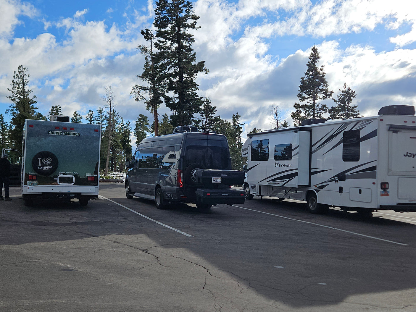 Three RVs, including a Cruise America rv rental and a Jayco Greyhawk, are parked outdoors amidst pine trees under a cloudy sky.
