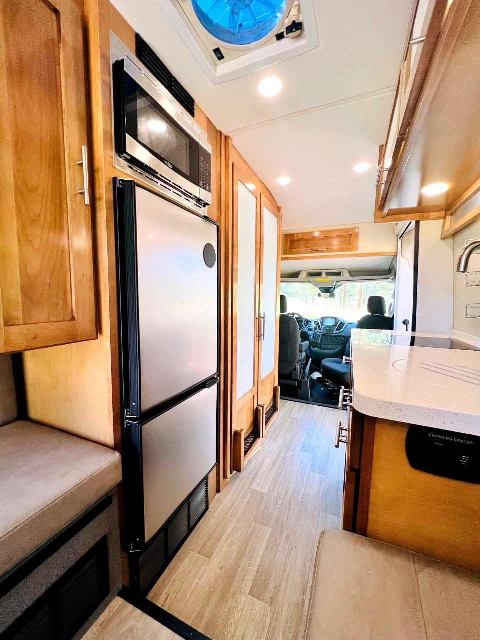 View inside a modern RV rental: a galley kitchen with microwave, fridge, sink, and light wood interior leading to the cab.