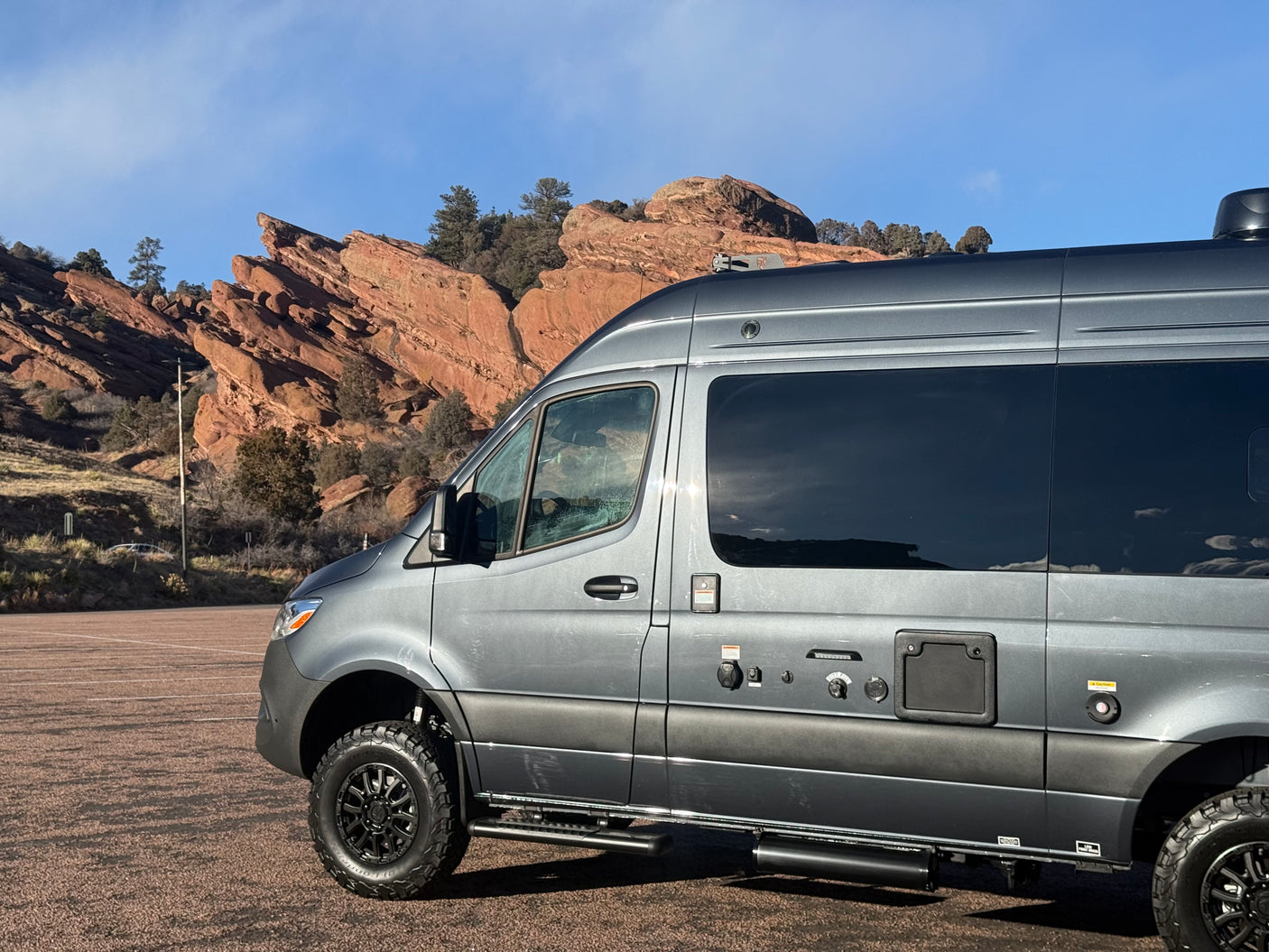 This grey RV rental van, equipped for adventure with large tires, is parked by red rock formations under a blue sky.