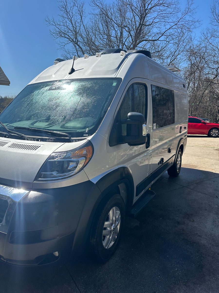 Silver camper van, ready for your RV rental journey, shines in the sun. Parked outdoors with bare trees and a red car in distance.