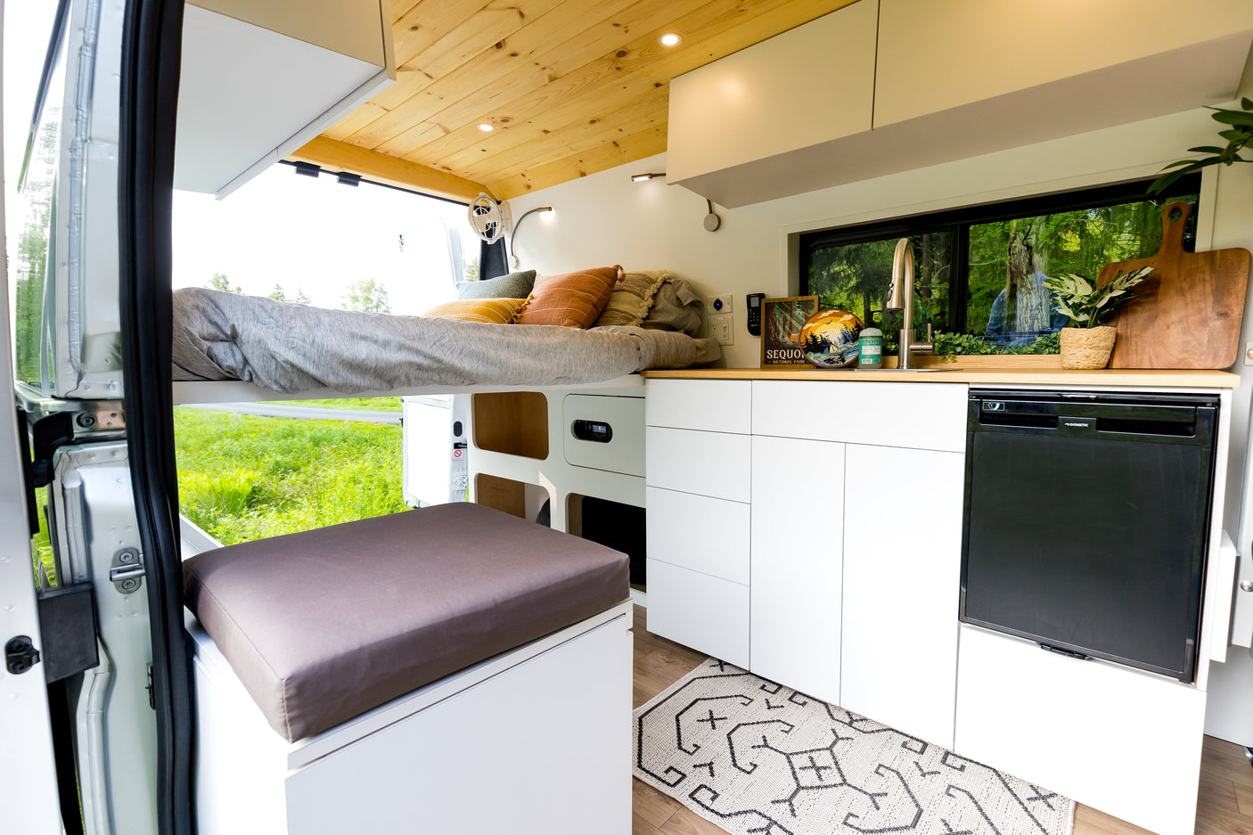 Cozy RV rental interior: raised bed, sleek kitchen with sink & fridge, white cabinets, and scenic window views.