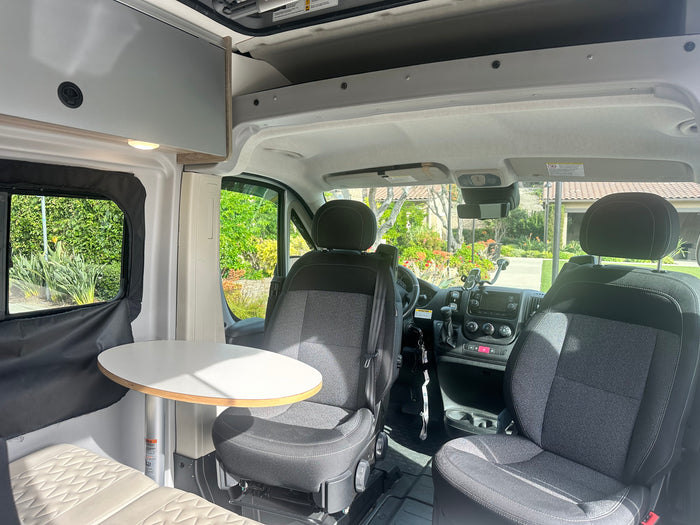 View of an RV rental camper van's interior: front seats, dashboard, dining table, & overhead storage.