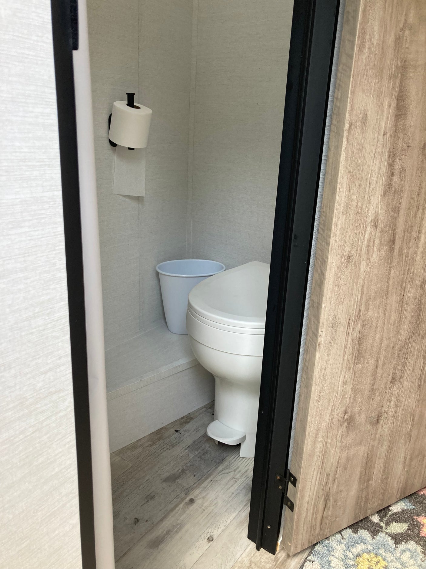 RV rental: A compact bathroom featuring a toilet, toilet paper roll, and white bucket on light wood flooring.