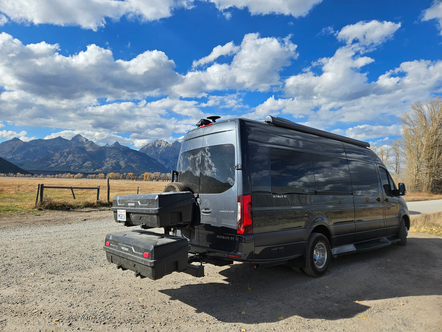 An RV rental van with rear storage, parked on a scenic dirt road, facing majestic mountains and a cloudy sky. Perfect for outdoor adventure.