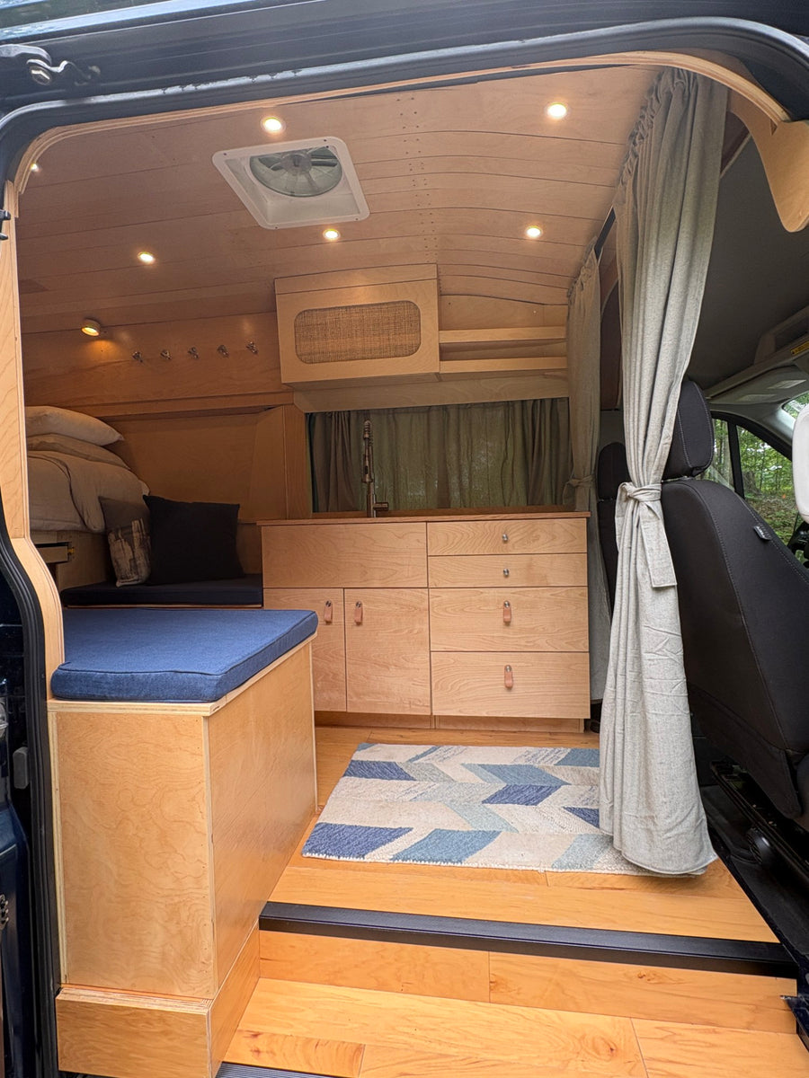 high-roof camper van parked on gravel in a forest. This type of vehicle is popular as an rv rental.|Explore this RV rental's cozy wood interior
