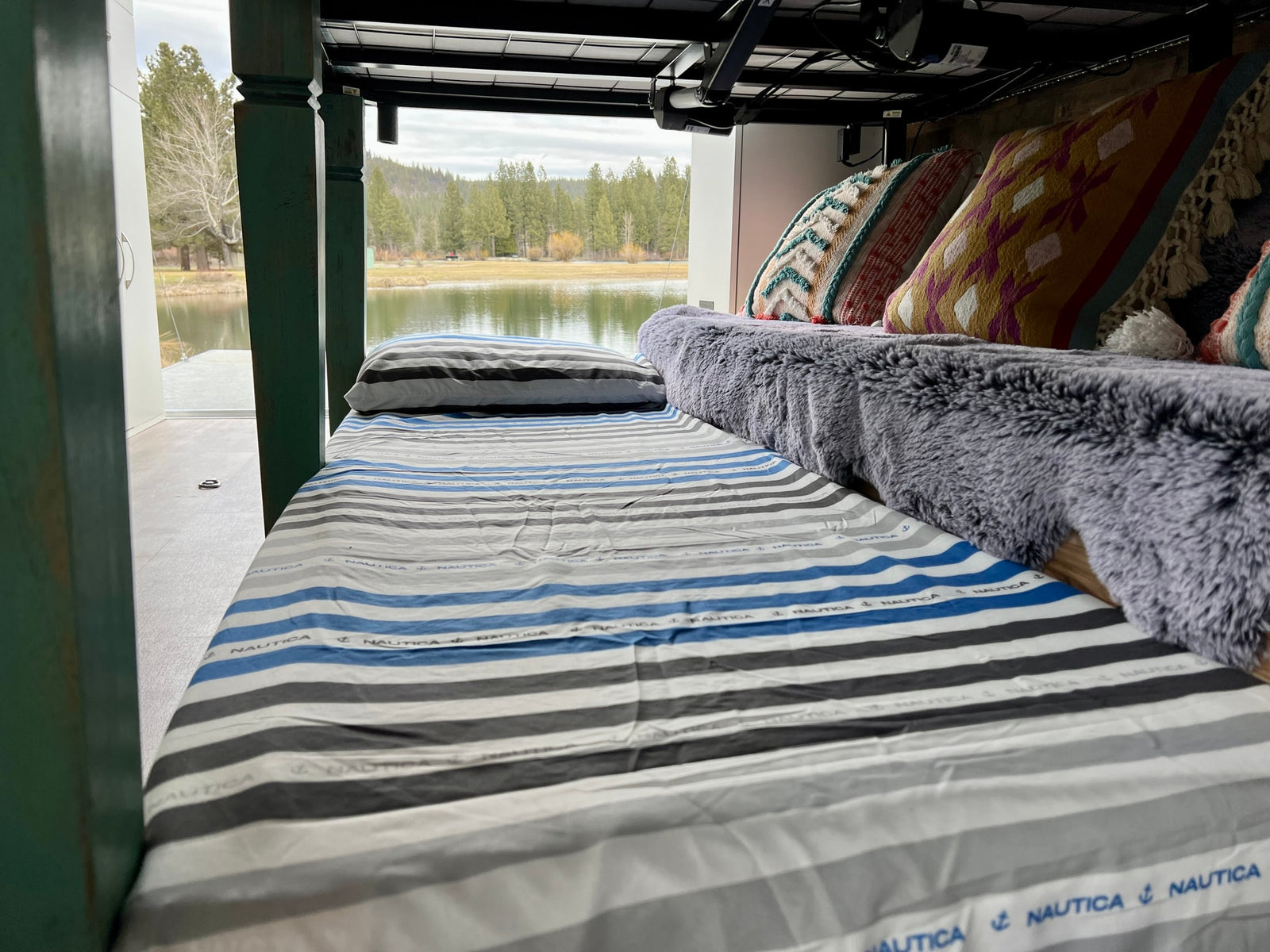 Experience tranquil travel from this RV rental bed with striped sheets, decorative pillows, and a scenic lake view.