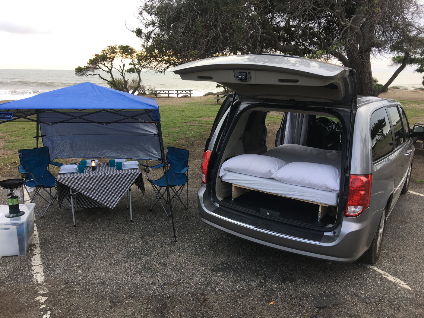 Minivan camper setup for beach camping, a great alternative to rv rental. Features a bed, canopy, & outdoor kitchen.