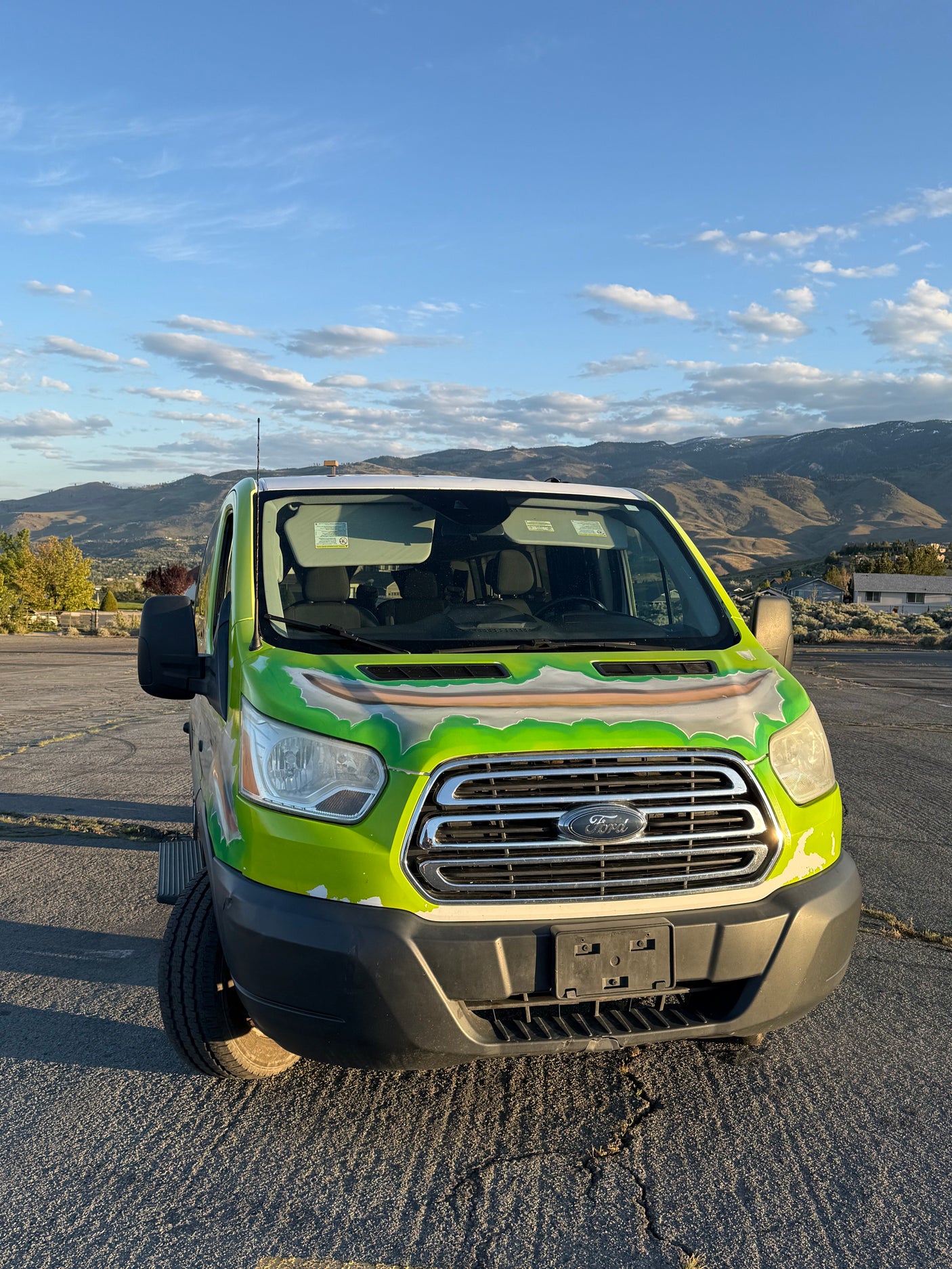 Front view of a bright green Ford Transit RV rental van, parked outdoors with mountains and a blue, cloudy sky.