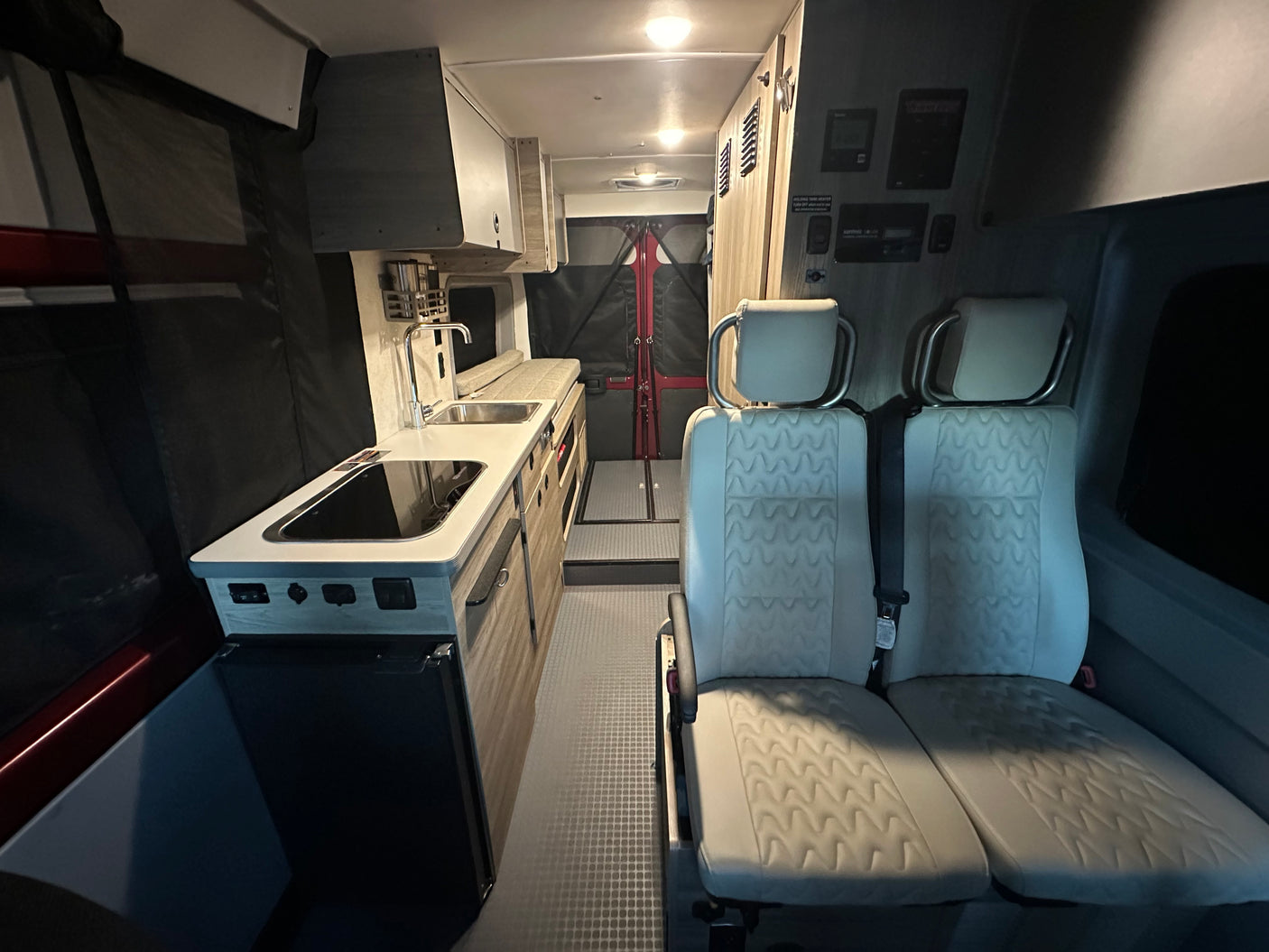 RV rental interior: A bright camper van featuring a compact kitchen with sink & stovetop, two patterned passenger seats, and overhead storage.