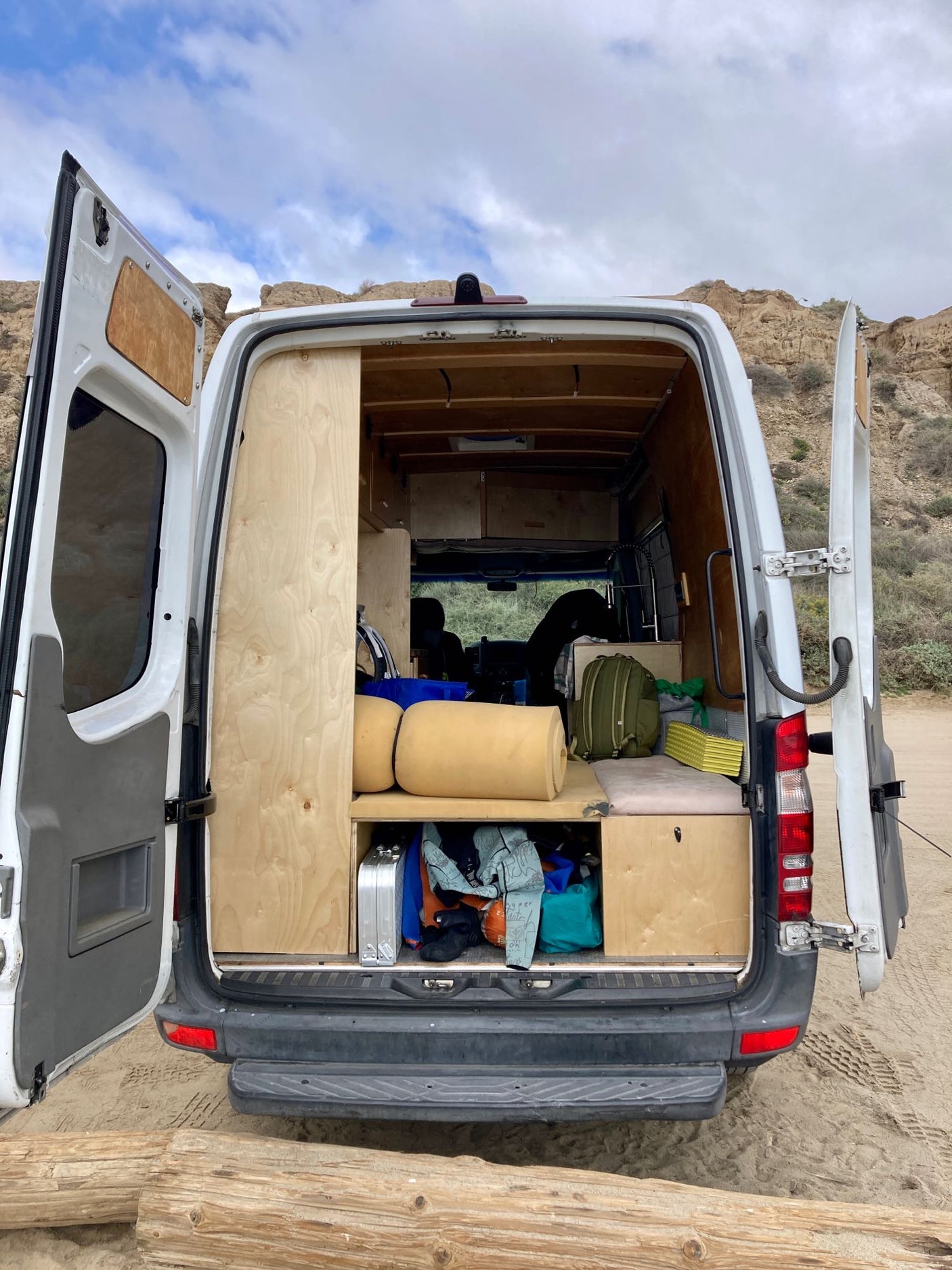 This converted van's open rear reveals a cozy interior & smart storage, perfect for an RV rental adventure on sandy terrain.