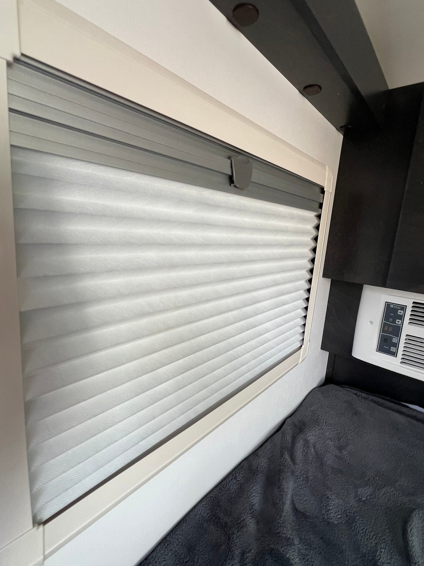 Cozy RV rental interior: window with pleated shade, climate control panel, and a soft blanket for comfort.