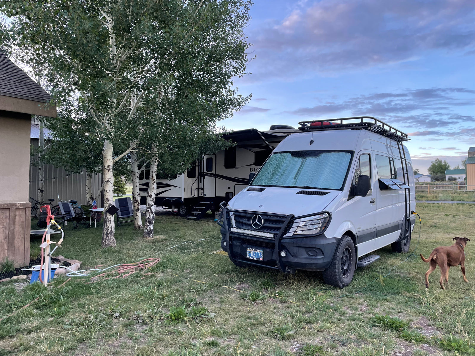 A white Mercedes Sprinter van RV rental and a large travel trailer set up in a grassy campsite with aspen trees and a dog.