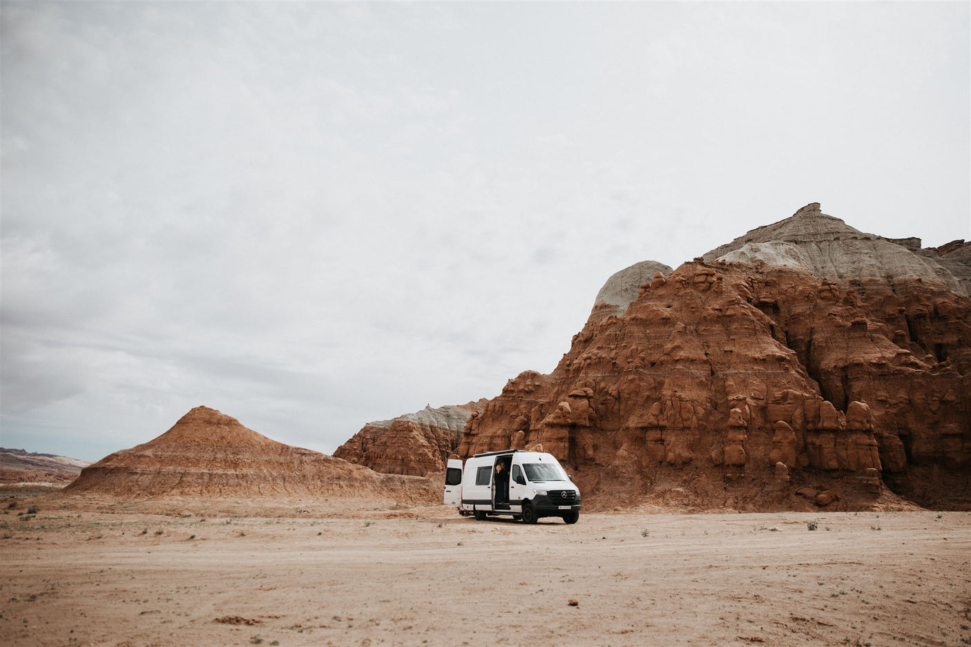 An RV rental in a dramatic desert landscape, parked amidst towering red rock formations under a cloudy sky.