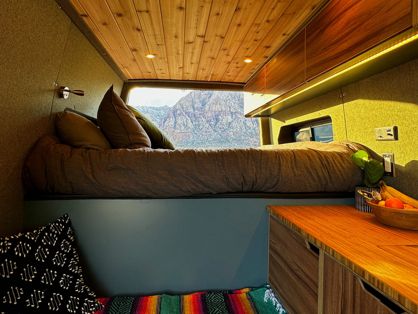 Relax in this RV rental's cozy interior, featuring a comfy bed, warm wood ceiling, and stunning mountain views from the back.
