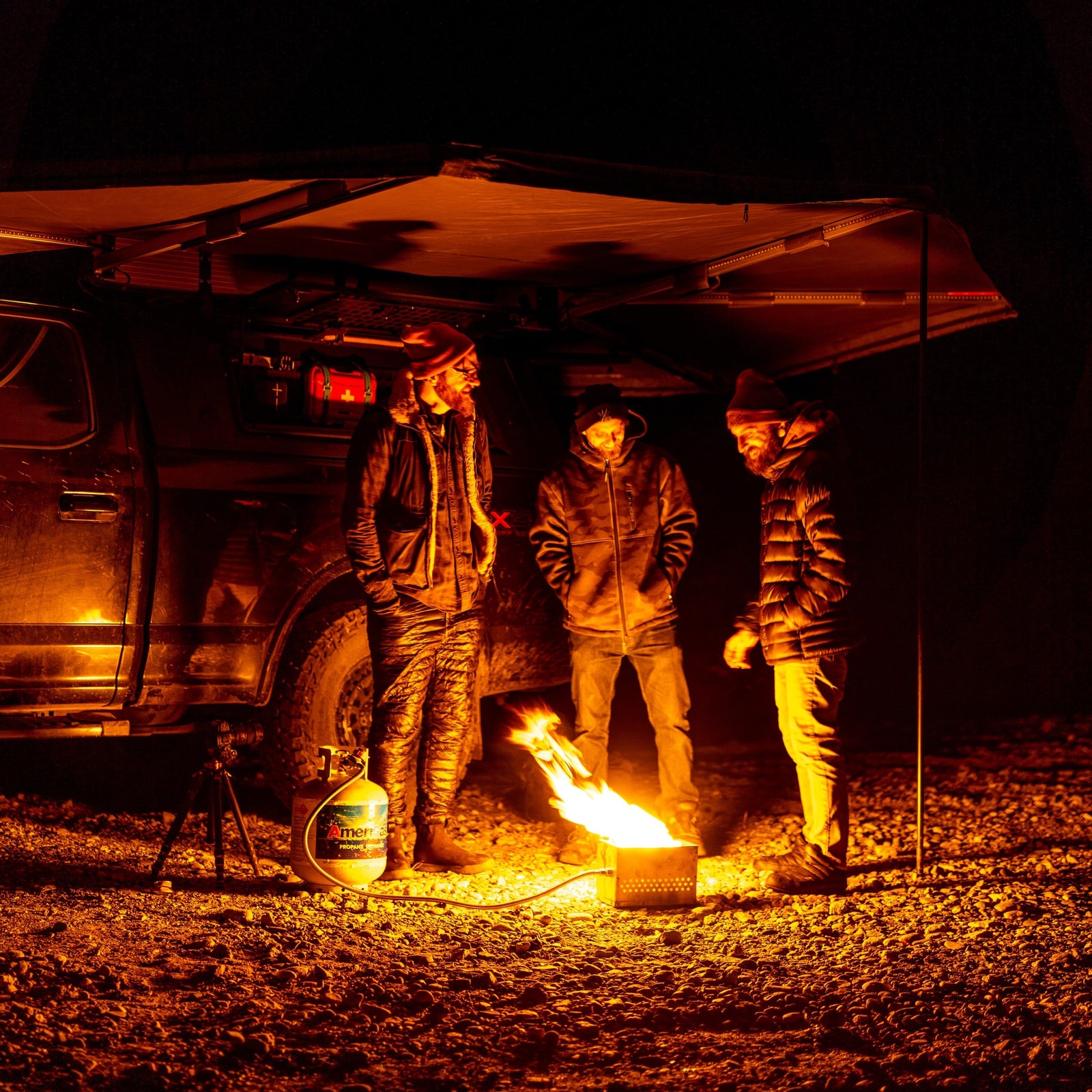 Night camping under an RV rental awning. Three men gathered around a cozy fire pit, enjoying the outdoors.