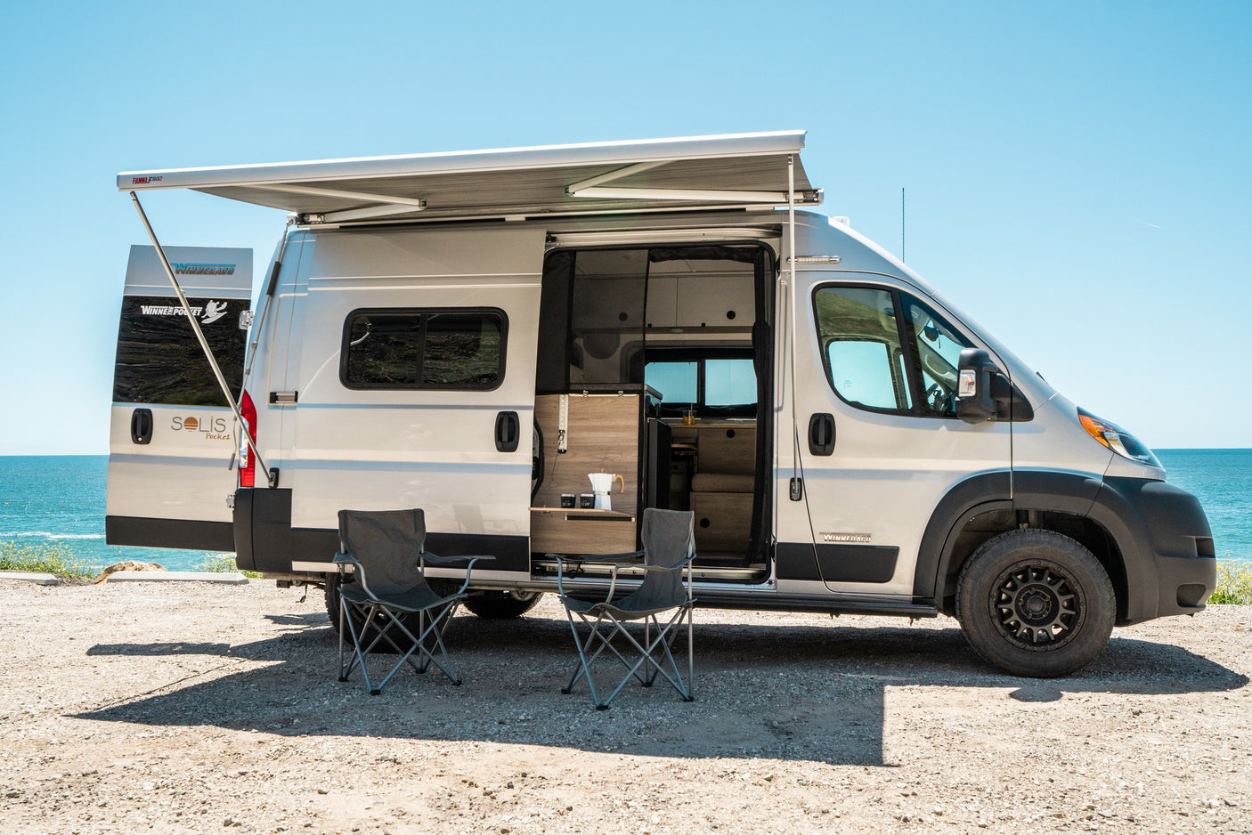 Coastal rv rental: Winnebago Solis Pocket camper van with awning, chairs & coffee setup by the sunny ocean.
