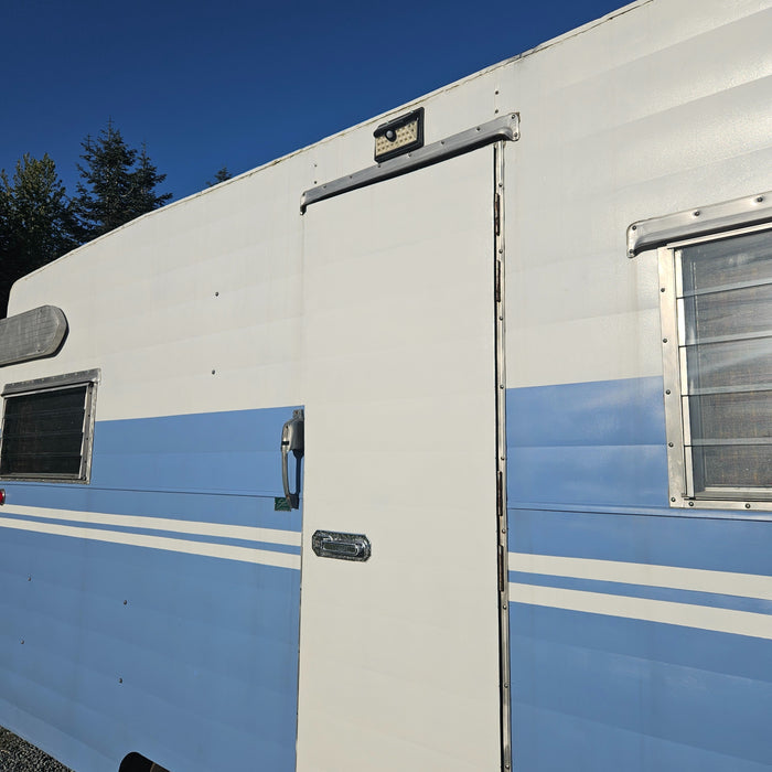 Close-up of a blue and white travel trailer's side, showing its entry door, windows, and a solar light. Consider this RV for rental.