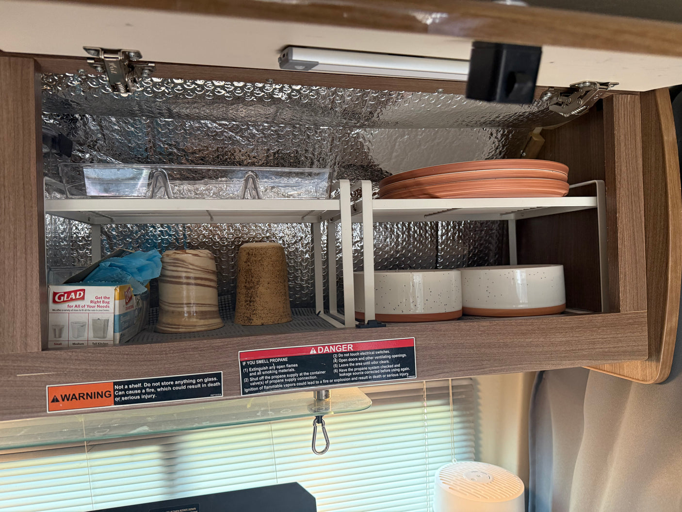 RV rental kitchen cabinet with organized shelves, plates, cups, and safety warnings, insulated with bubble wrap.