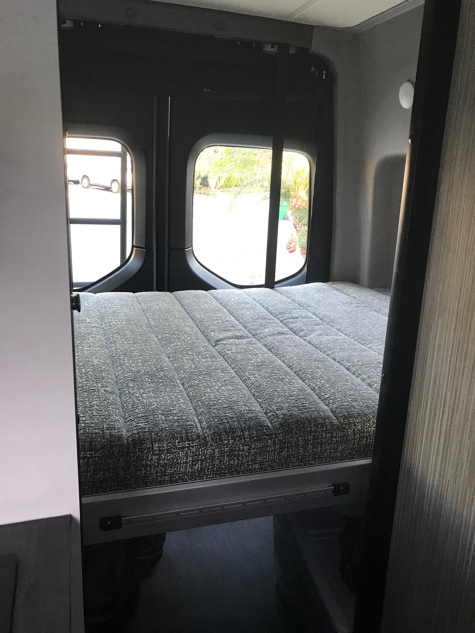 RV rental: Compact sleeping area with a grey textured bed and rear windows, inside a camper van.