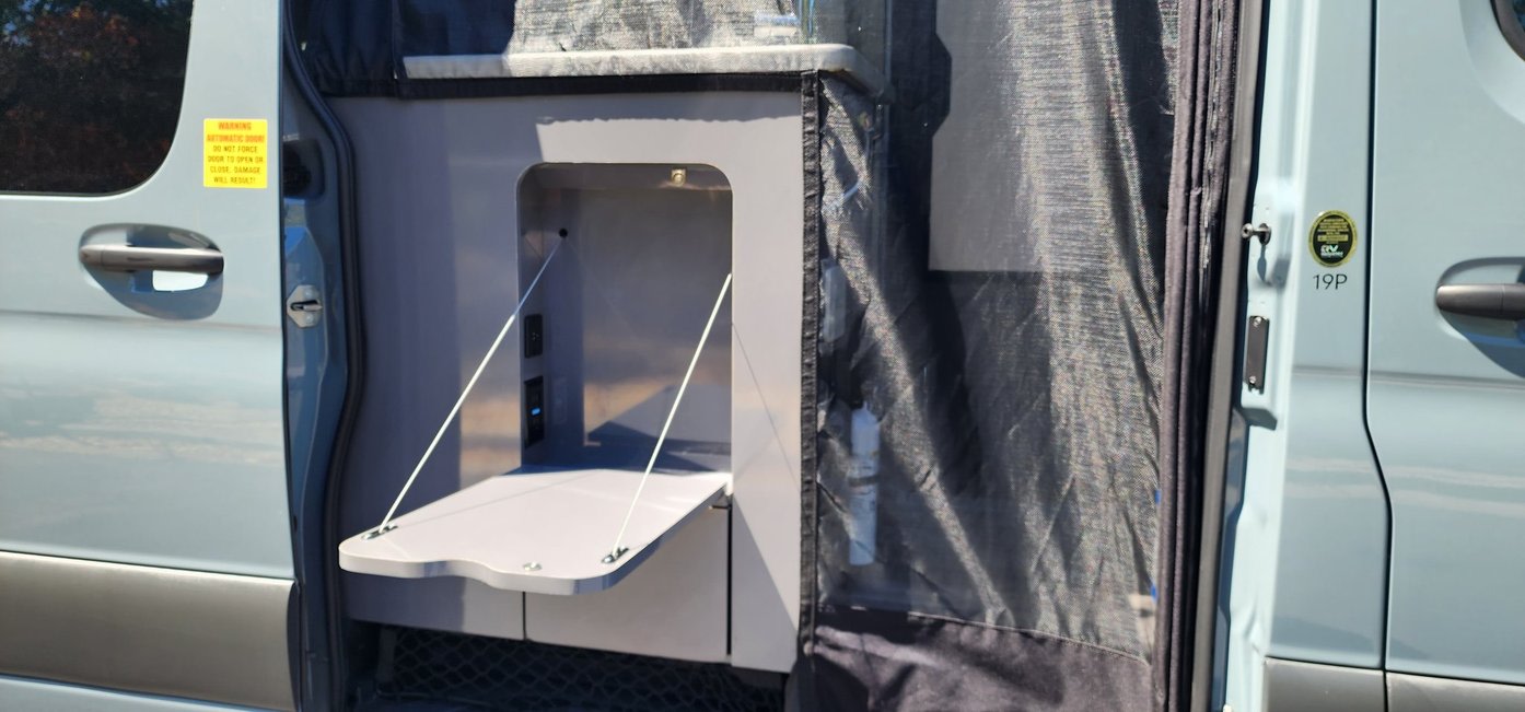 Interior of an RV rental van, featuring an open side door with a fold-down table and power outlets.