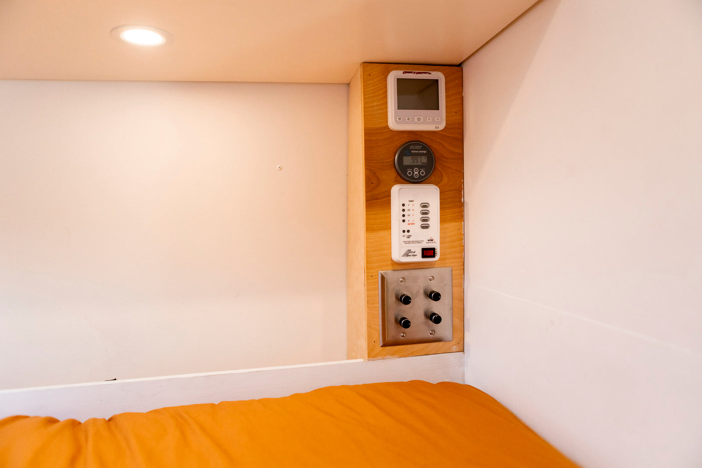 RV rental: Close-up of interior controls (Victron, tank/battery monitor, thermostat) above a comfy orange bed.