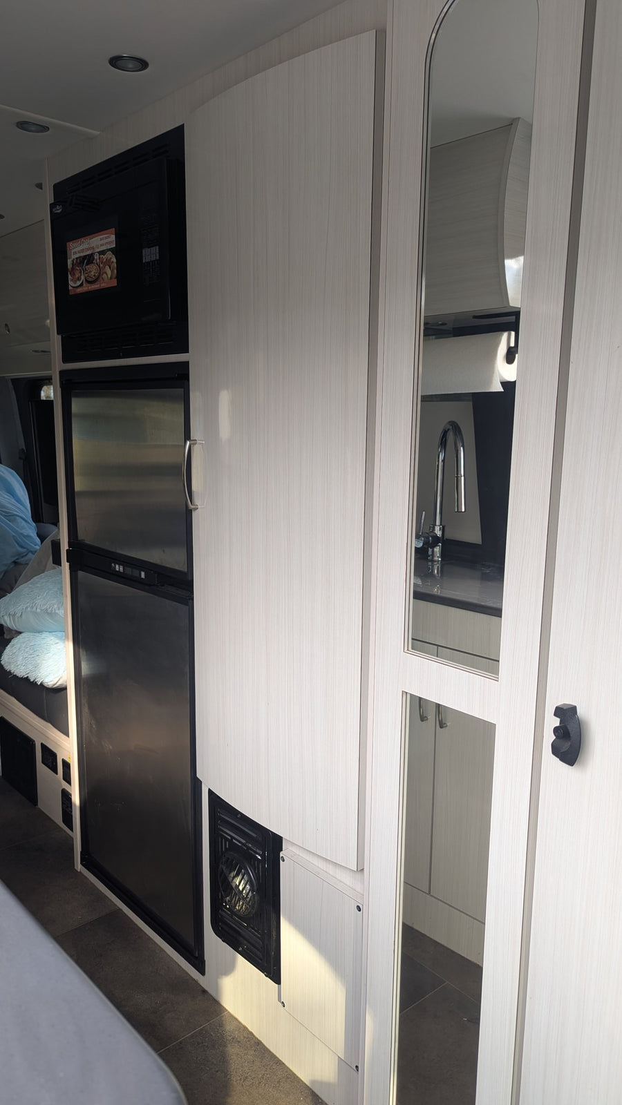 This RV rental offers a compact kitchen with microwave, fridge, and light wood cabinets. Mirrored doors reflect the sink area.