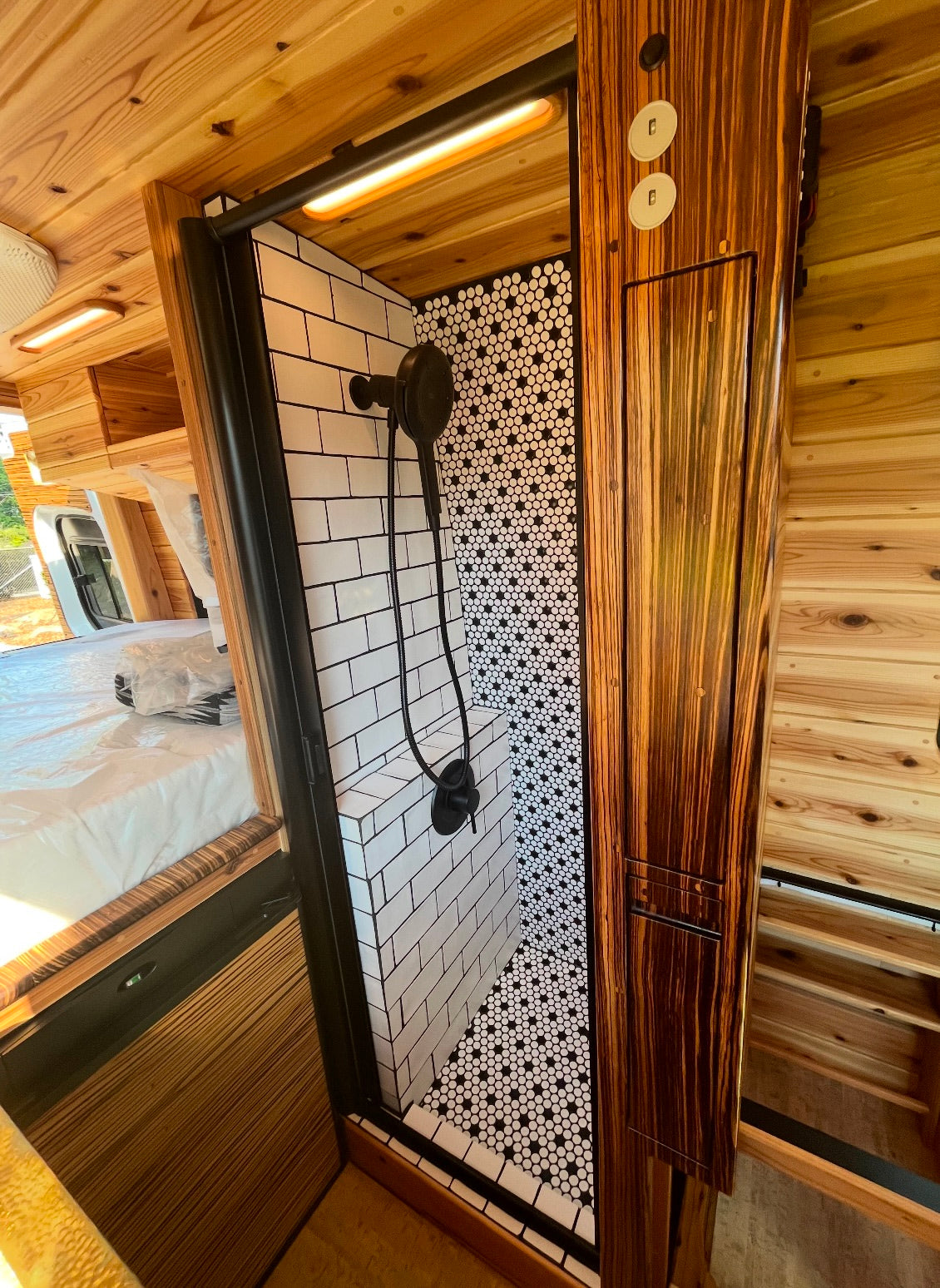 Modern RV rental shower with subway tile walls, black & white hexagon floor, sleek black fixtures, and natural wood interior.