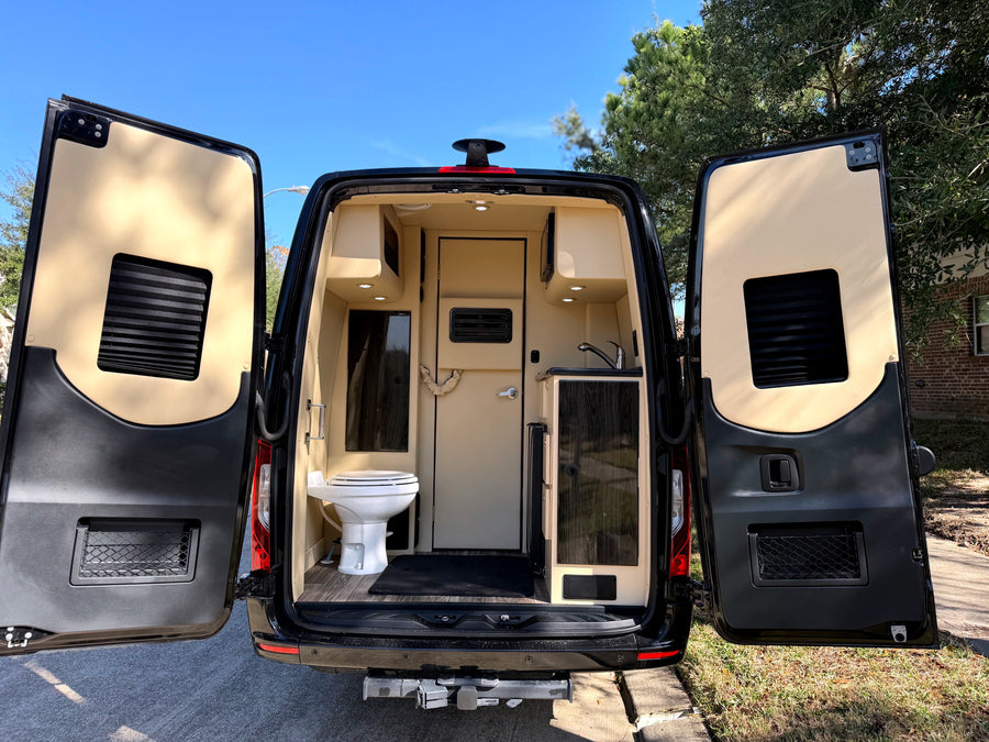 This rv rental's open rear doors reveal a functional interior: toilet, sink, and tan cabinets under bright lights.