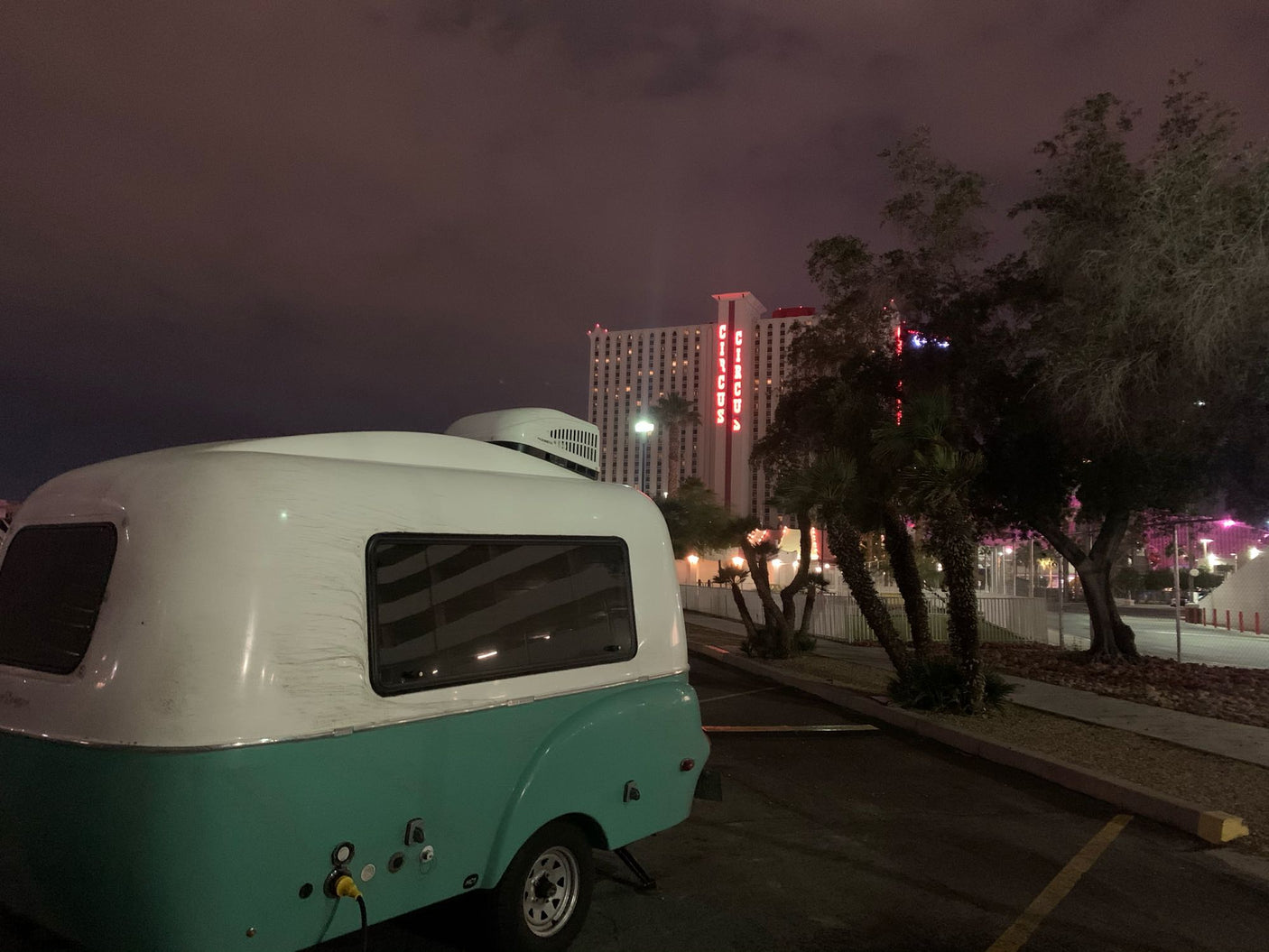 Night view of a retro **rv rental** camper, white & turquoise, parked at Circus Circus Las Vegas. Bright neon sign and trees visible.