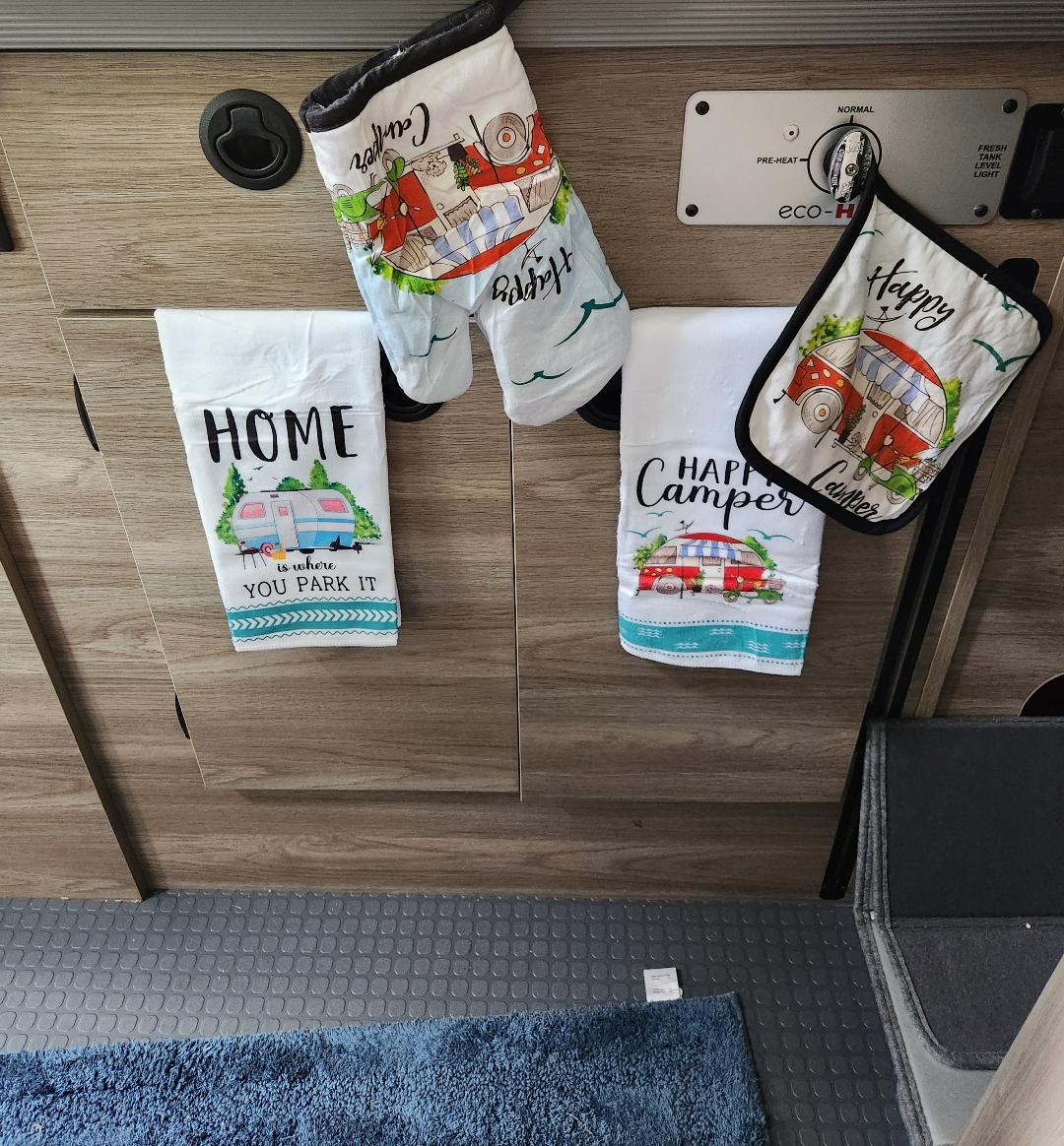 Inside an RV rental: fun camper-themed towels & oven mitts hang on wooden cabinets, adding a cozy camping vibe.