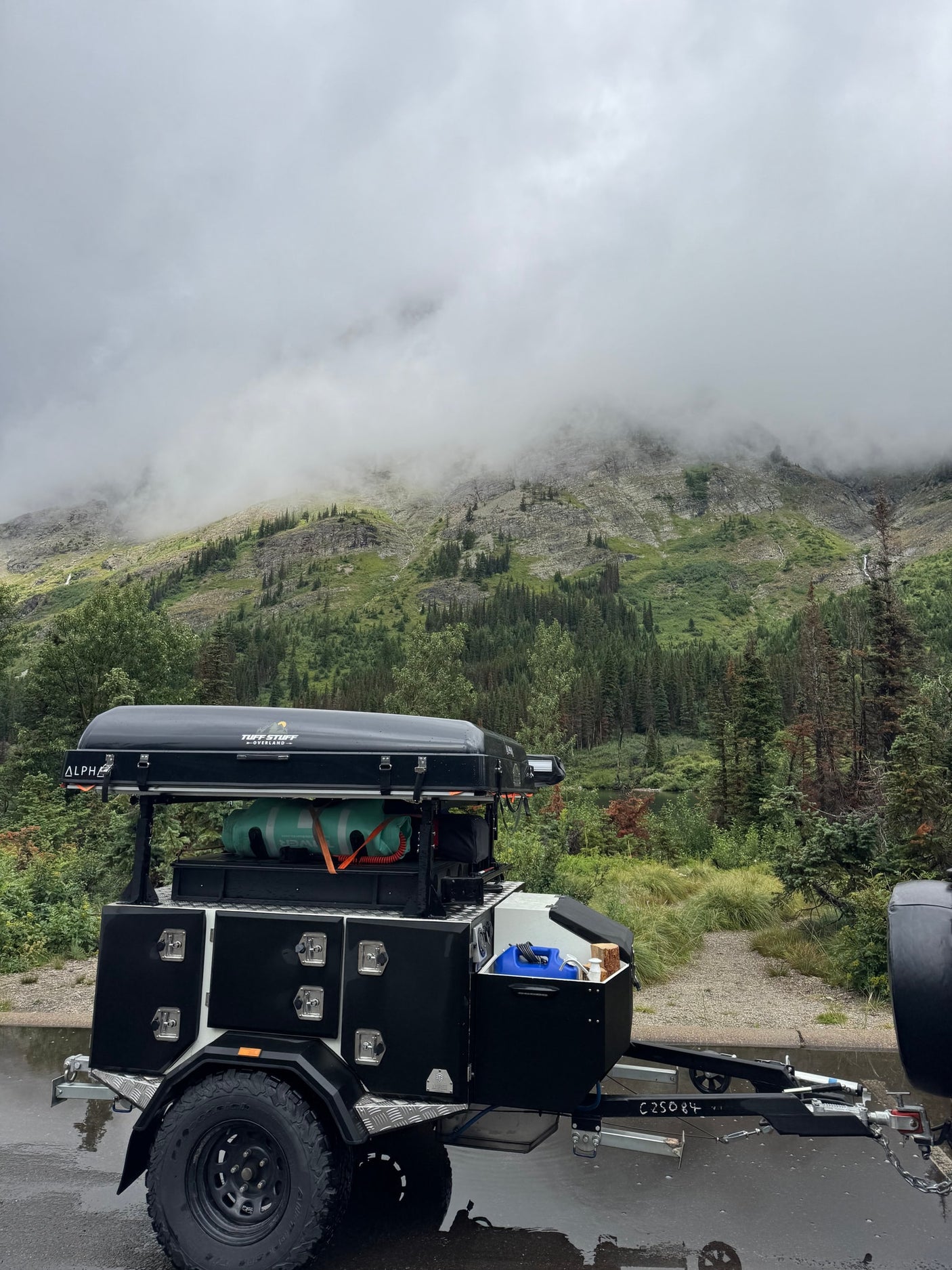 Overland trailer with rooftop tent on a wet road, misty mountains in the background. Ideal for your next RV rental adventure.