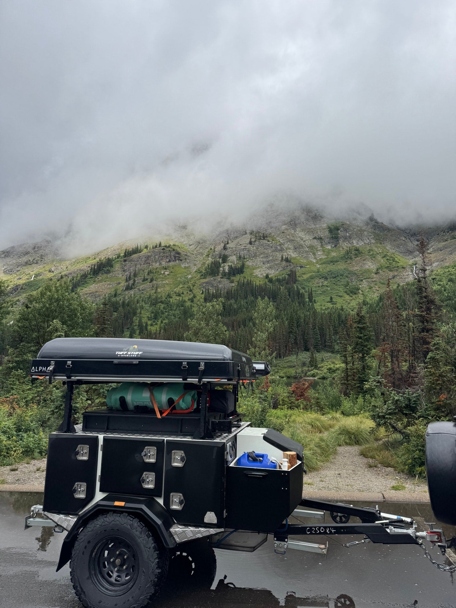 Overland trailer with rooftop tent on a wet road, misty mountains in the background. Ideal for your next RV rental adventure.