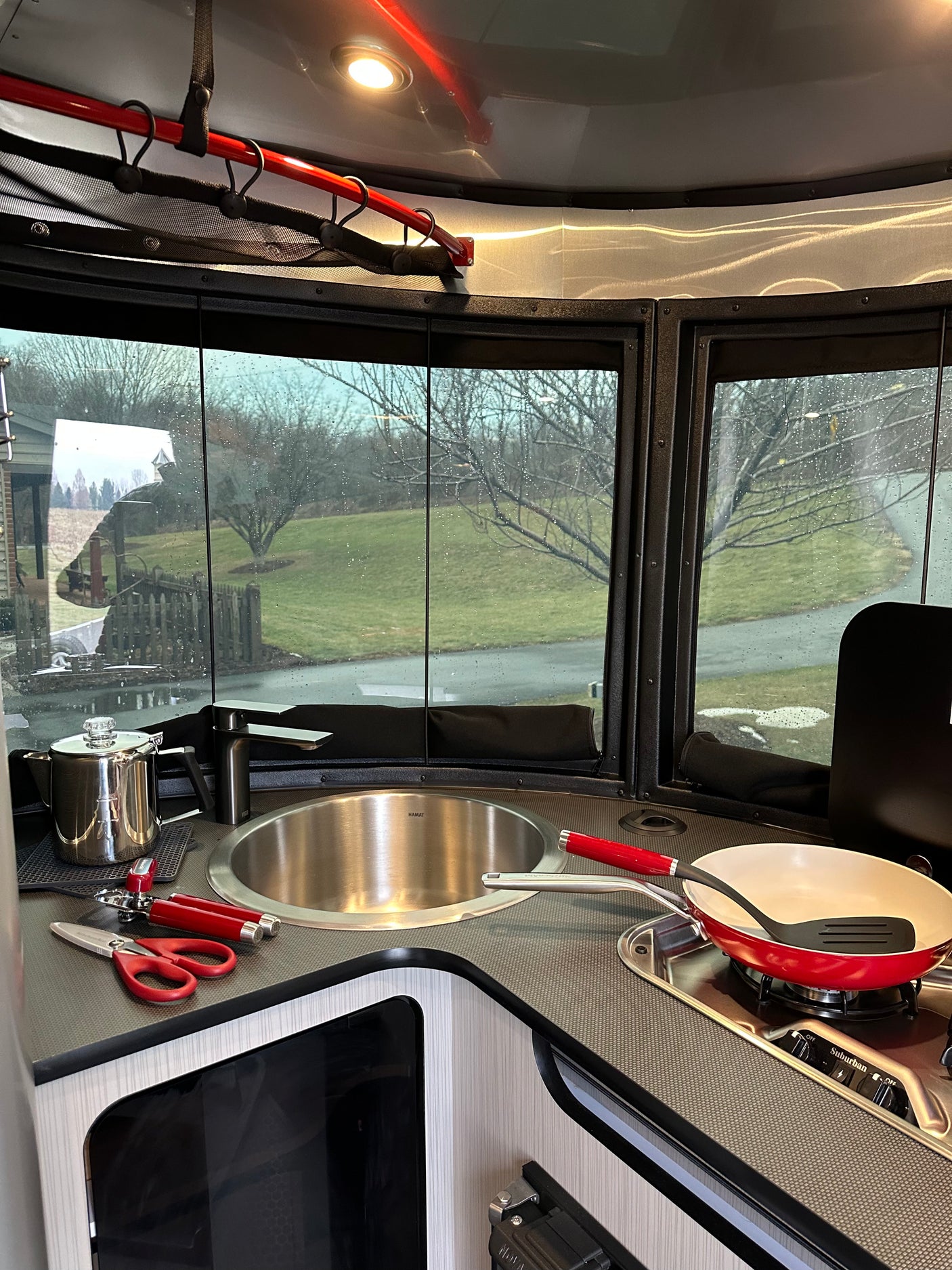 Modern RV rental kitchen with sink, stovetop, and red cookware. Large windows offer a view of a grassy, treed landscape.