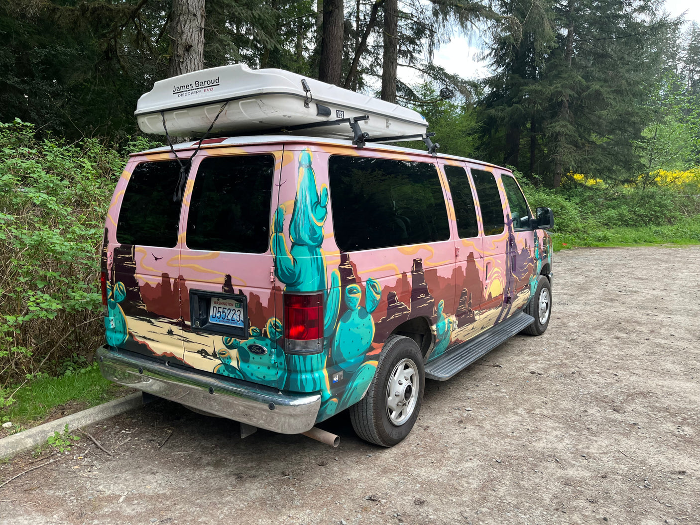 This customized rv rental, painted with a desert mural, features a rooftop tent and is parked in a forest setting.