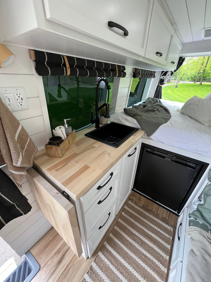 RV rental: Modern camper van interior shows a compact kitchen with wood counter, black sink/faucet, fridge, and bed.