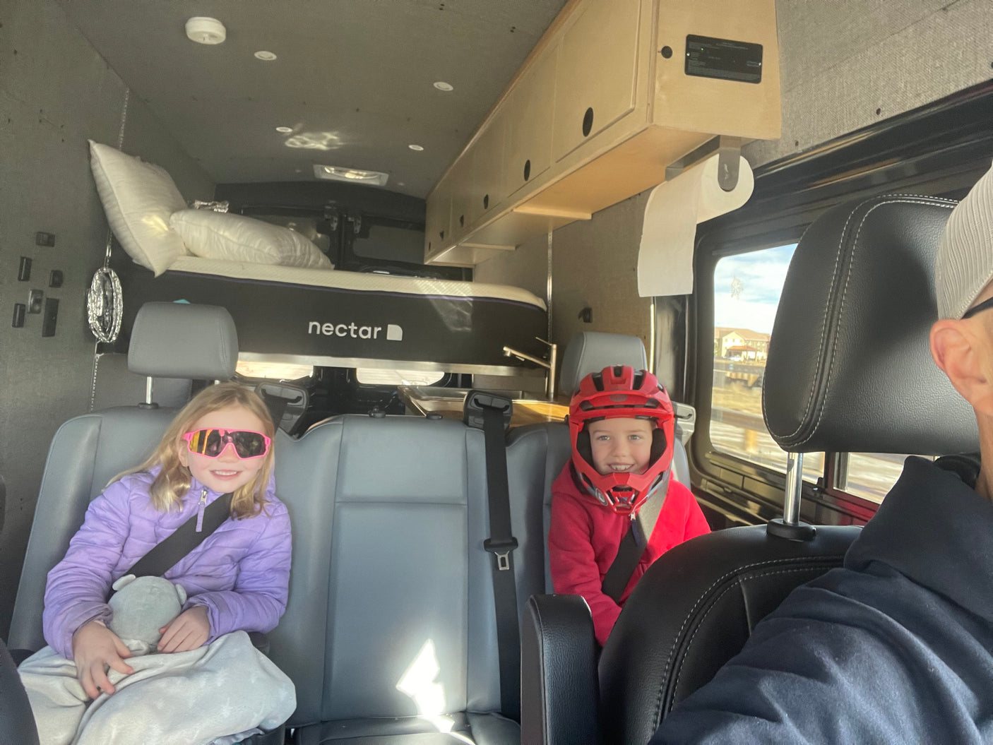 Happy children seated in an RV rental, showcasing the camper van's custom interior, bunk bed with Nectar mattress, and overhead storage.