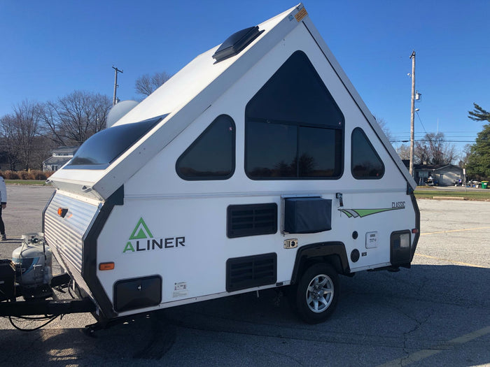 surrounded by pine trees.|A white A-Liner A-frame travel trailer