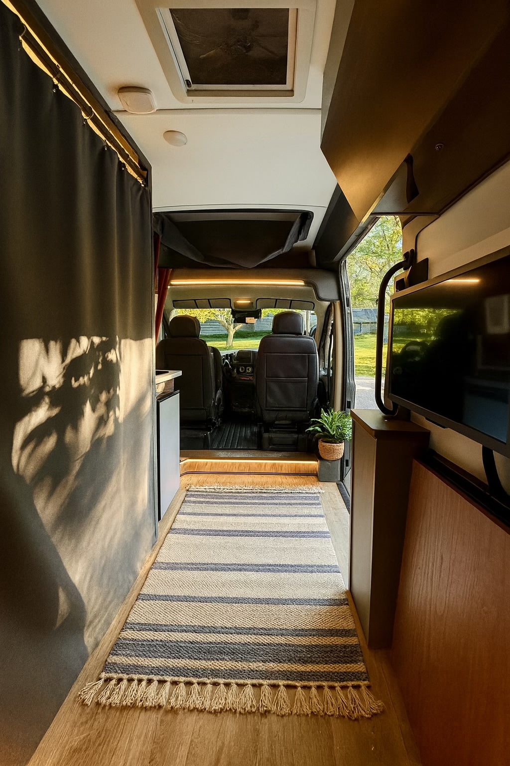 Rv rental: Cozy camper van interior featuring wooden floor, striped rug, accent lighting, and a TV, looking towards the cab area.