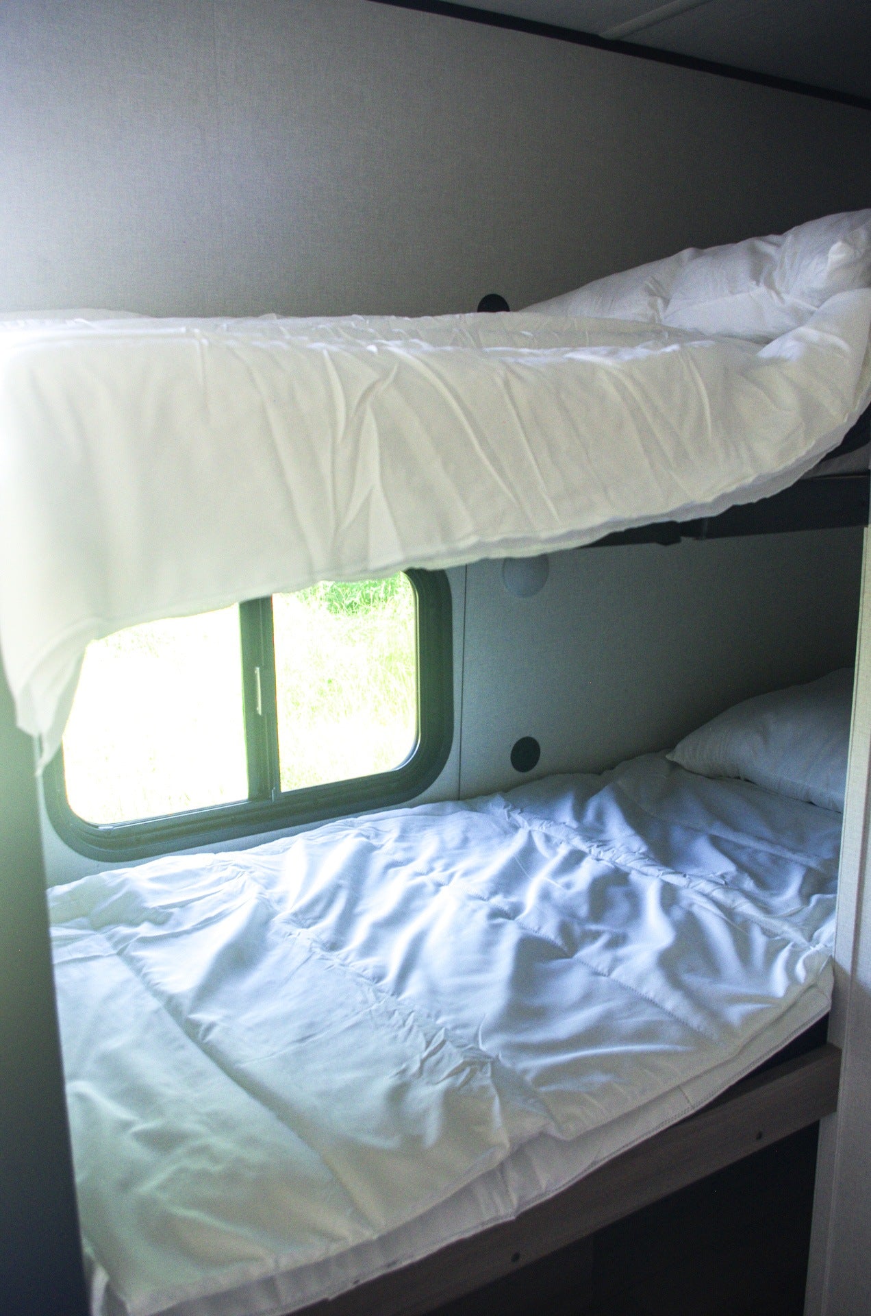 Clean, cozy bunk beds with white linens in an RV rental. A window reveals green nature outside, ideal for travel.