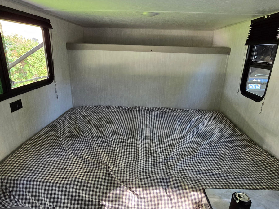 Inside an RV rental: a compact sleeping area with a checkered bed, overhead shelf, and windows showing nature & a Jeep.